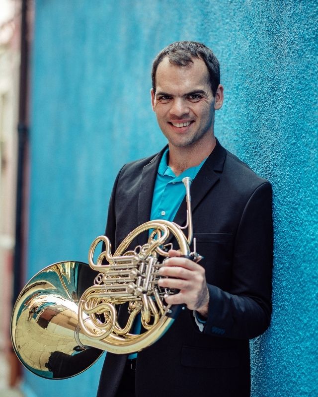 Matthew Mauro, Anthony Wayne Class of 2007, smiling while holding a French horn against a bright blue wall.